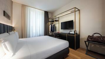 A hotel room with a bed, a desk, a chair, a TV, a coat rack, and a handbag.