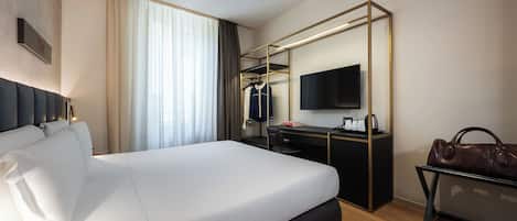 A hotel room with a bed, a desk, a chair, a TV, a coat rack, and a handbag.