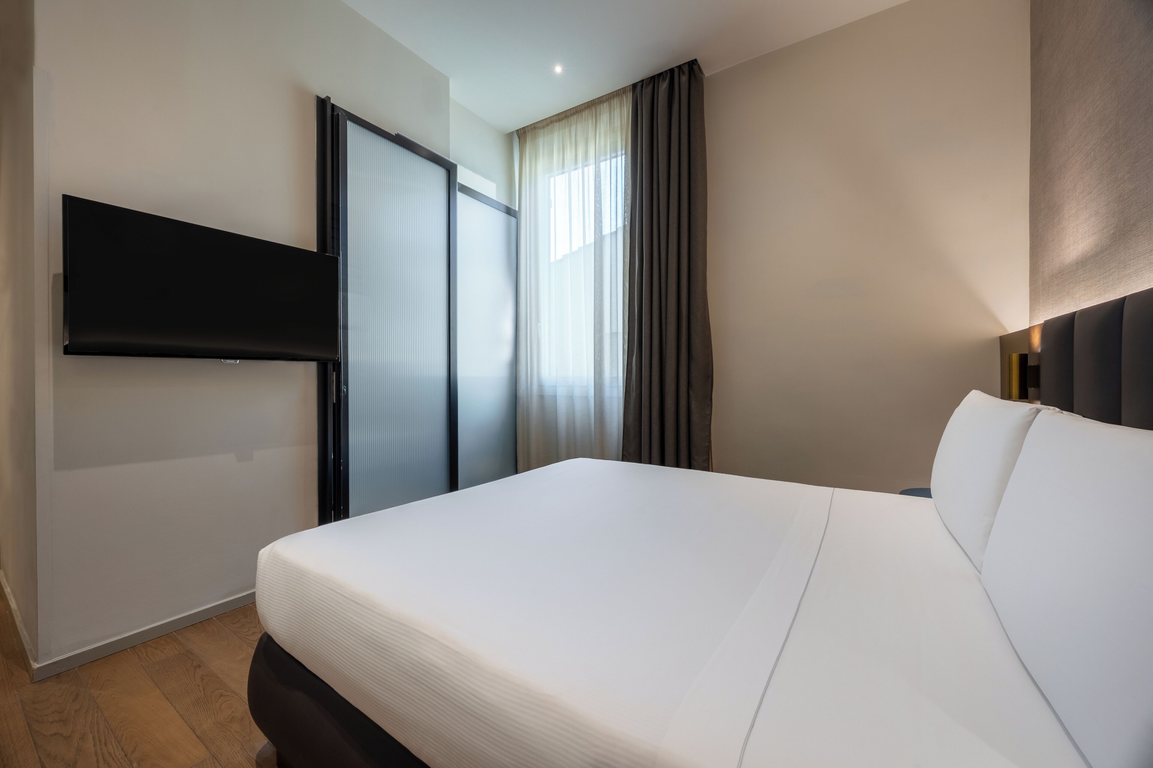 A hotel room with a large bed, a flat-screen TV mounted on the wall, a sliding door, and a window with curtains.