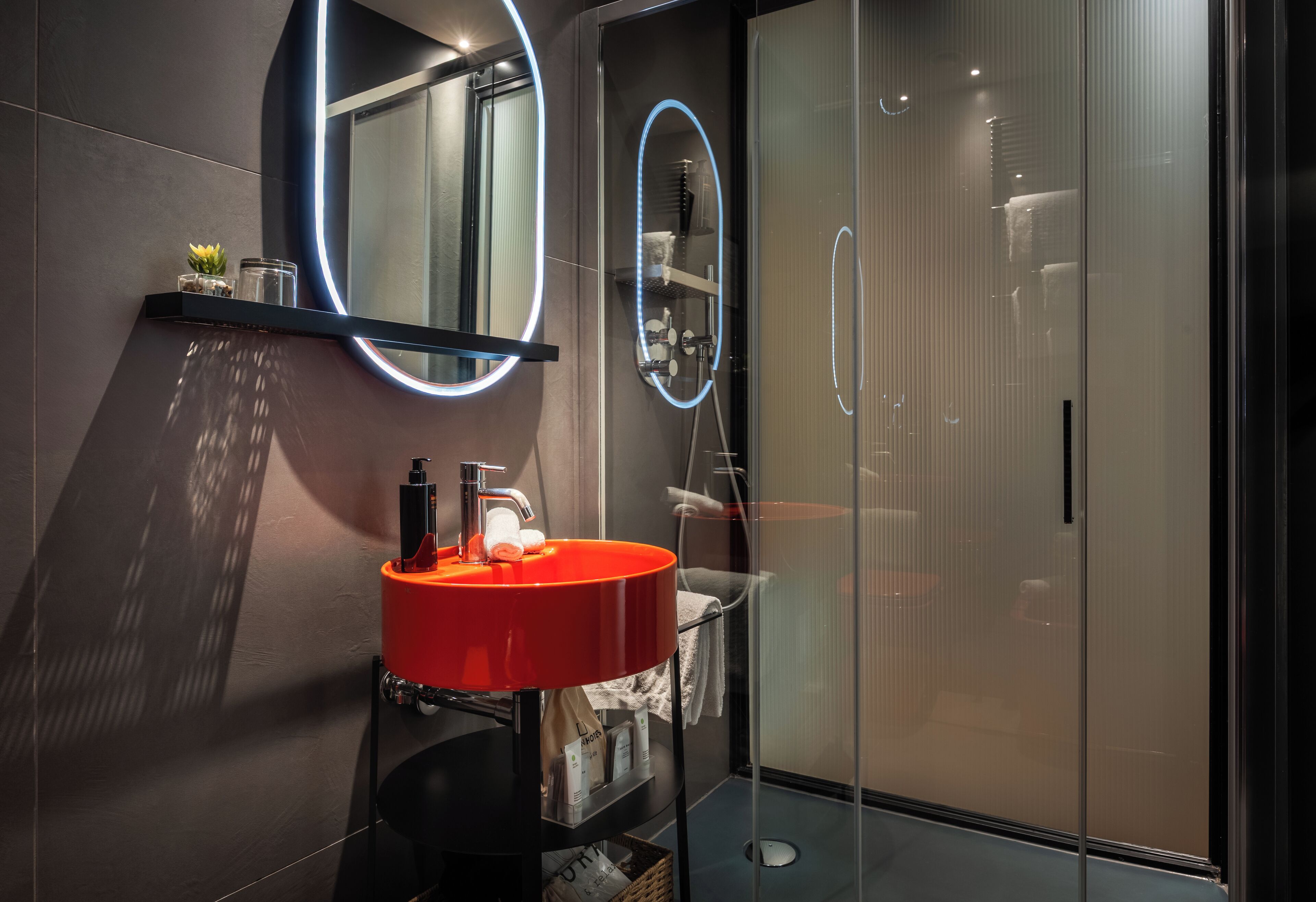 Un baño moderno con un lavabo rojo, un espejo grande con luz de neón y una cabina de ducha de vidrio.