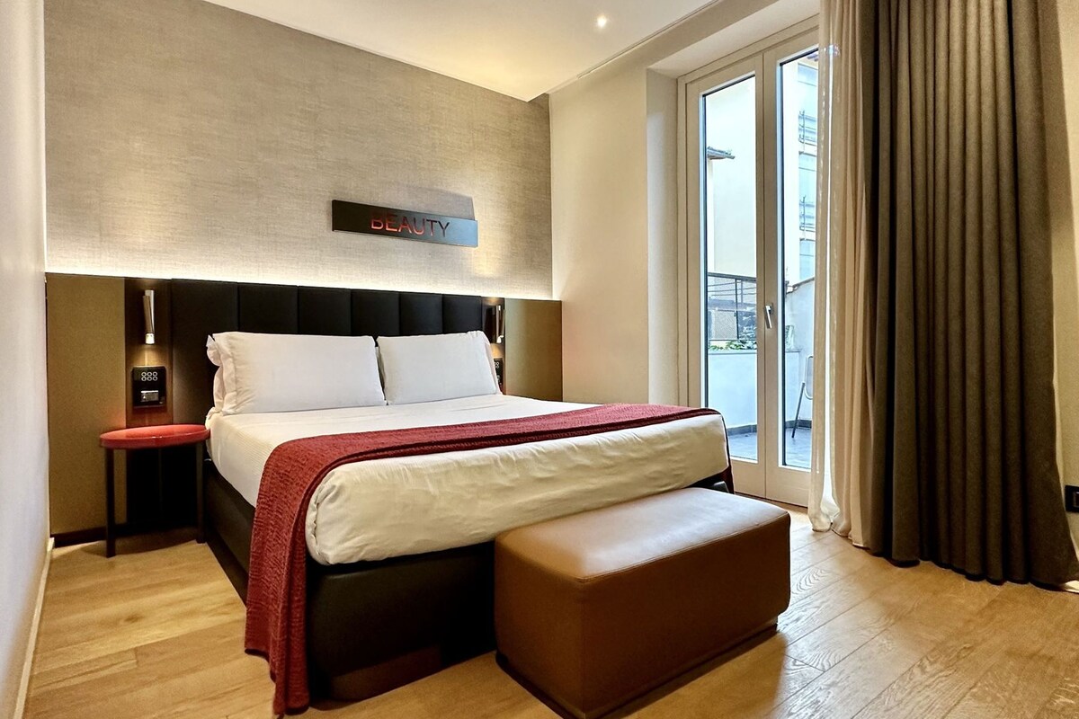 A hotel room with a large bed, a bedside table, a red blanket, a bench, a wall-mounted control panel, and a view through a sliding door.