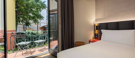 Habitación de hotel con una cama grande, una mesita de noche con lámpara y un ventanal corredero que da a un área exterior con mesa y sillas.