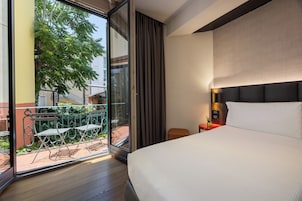 A hotel room with a large bed, a nightstand with a lamp, and a sliding glass door leading to an outdoor area with a table and chairs.