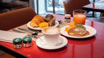 Une table rouge dressée pour le petit-déjeuner, avec des croissants, des muffins, des crêpes et une tasse de café.