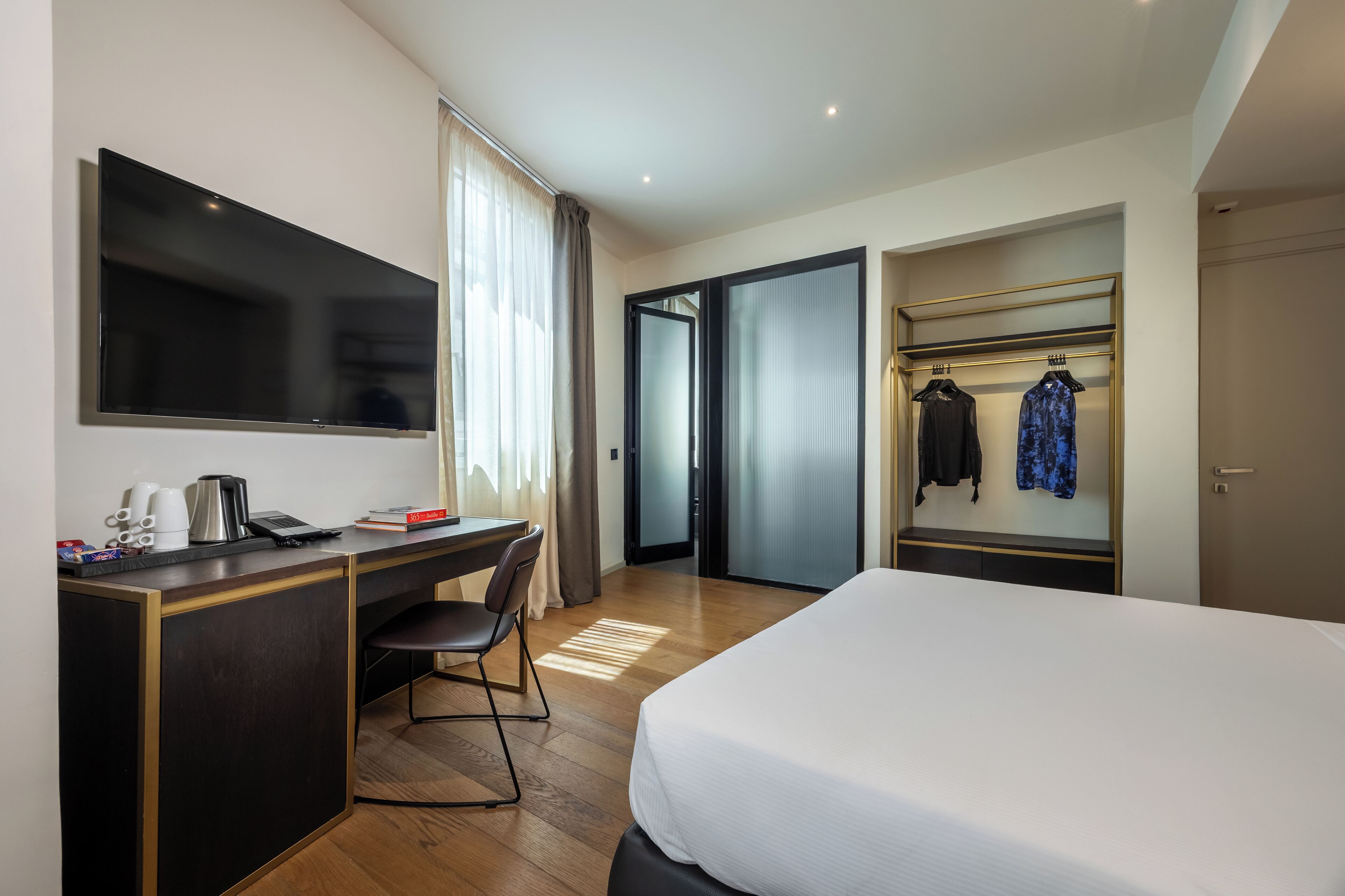 A hotel room with a bed, desk, chair, TV, and a wardrobe with clothes.