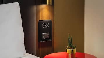 Quarto de hotel com uma cama, uma mesa de cabeceira com tampo vermelho, uma pequena planta em vaso e um painel de controle fixado na parede.