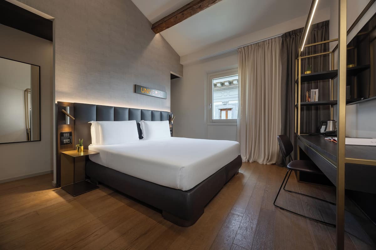 A modern hotel room with a large bed, bedside tables, a desk, and a bookshelf.