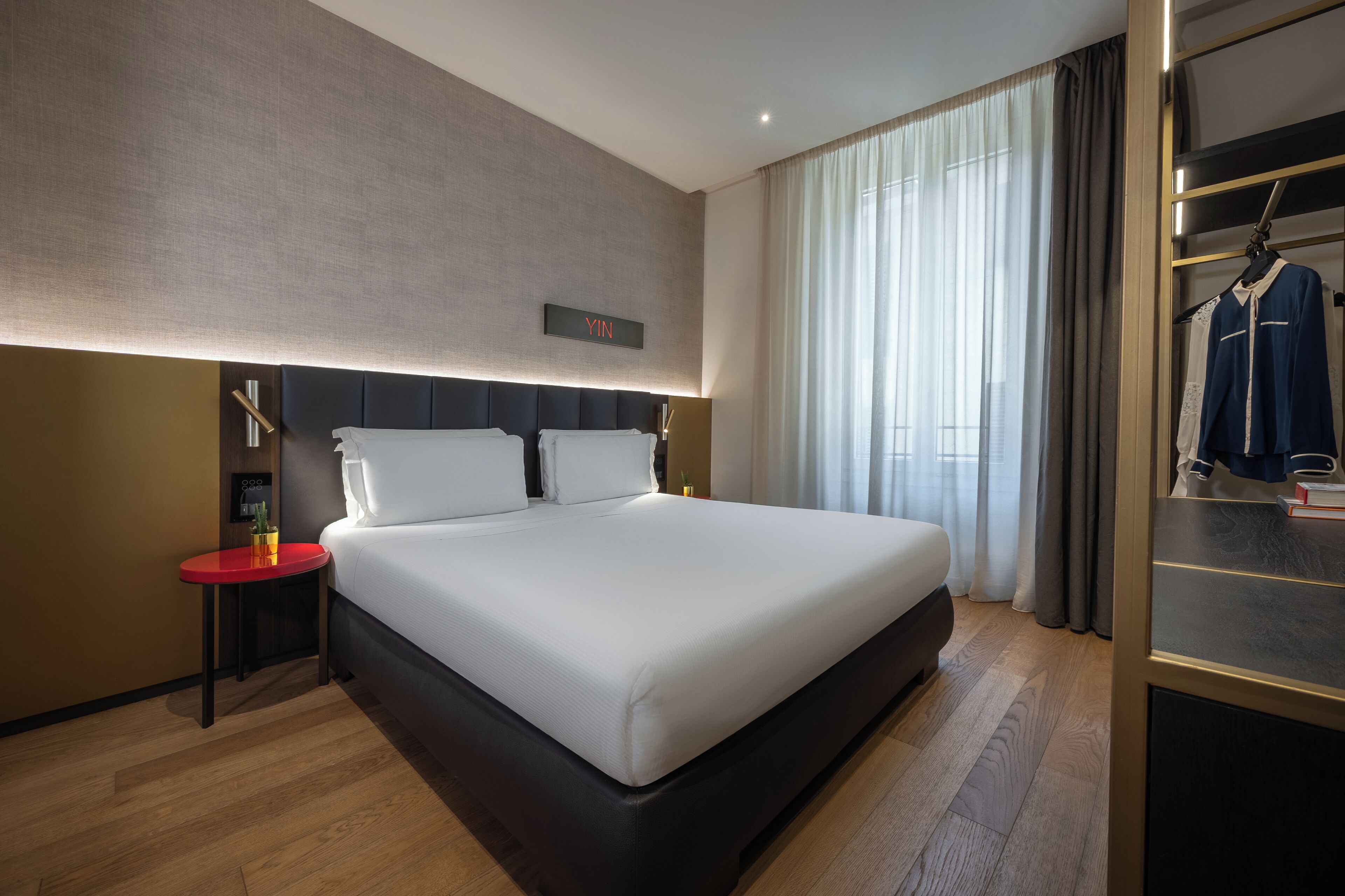 A modern hotel room with a large bed, a bedside table with a lamp, a wardrobe, and a sign with the name 'YIN'.