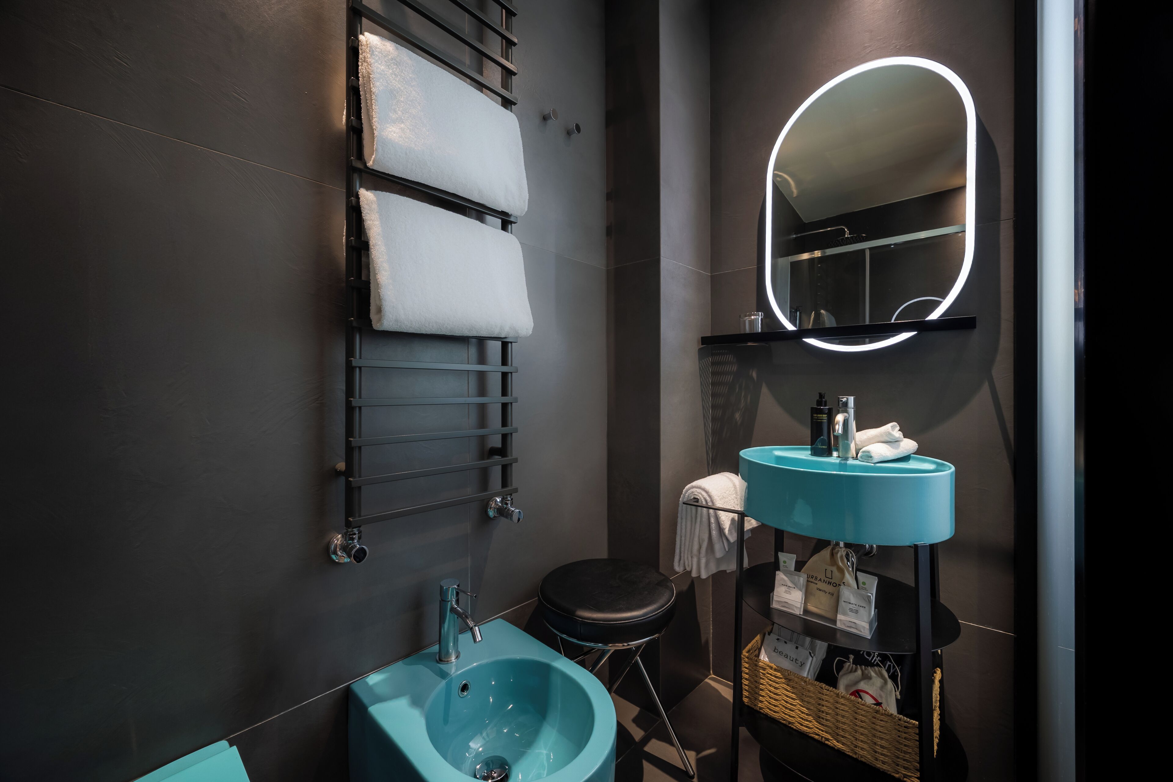 Un baño moderno con un lavamanos verde azulado, un espejo con luz de neón y un perchero con toallas dobladas.