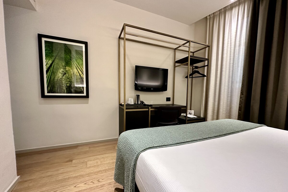 A hotel room with a bed, a desk with a chair, a television, and a framed picture on the wall.