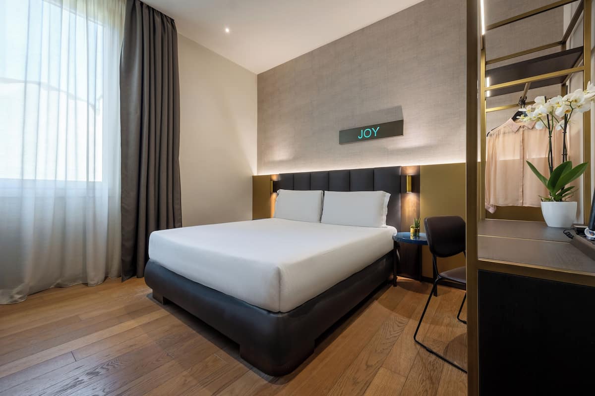 A modern hotel room with a large bed, a bedside table, a desk with a chair, a mirror, and a sign that says "JOY" on the wall.