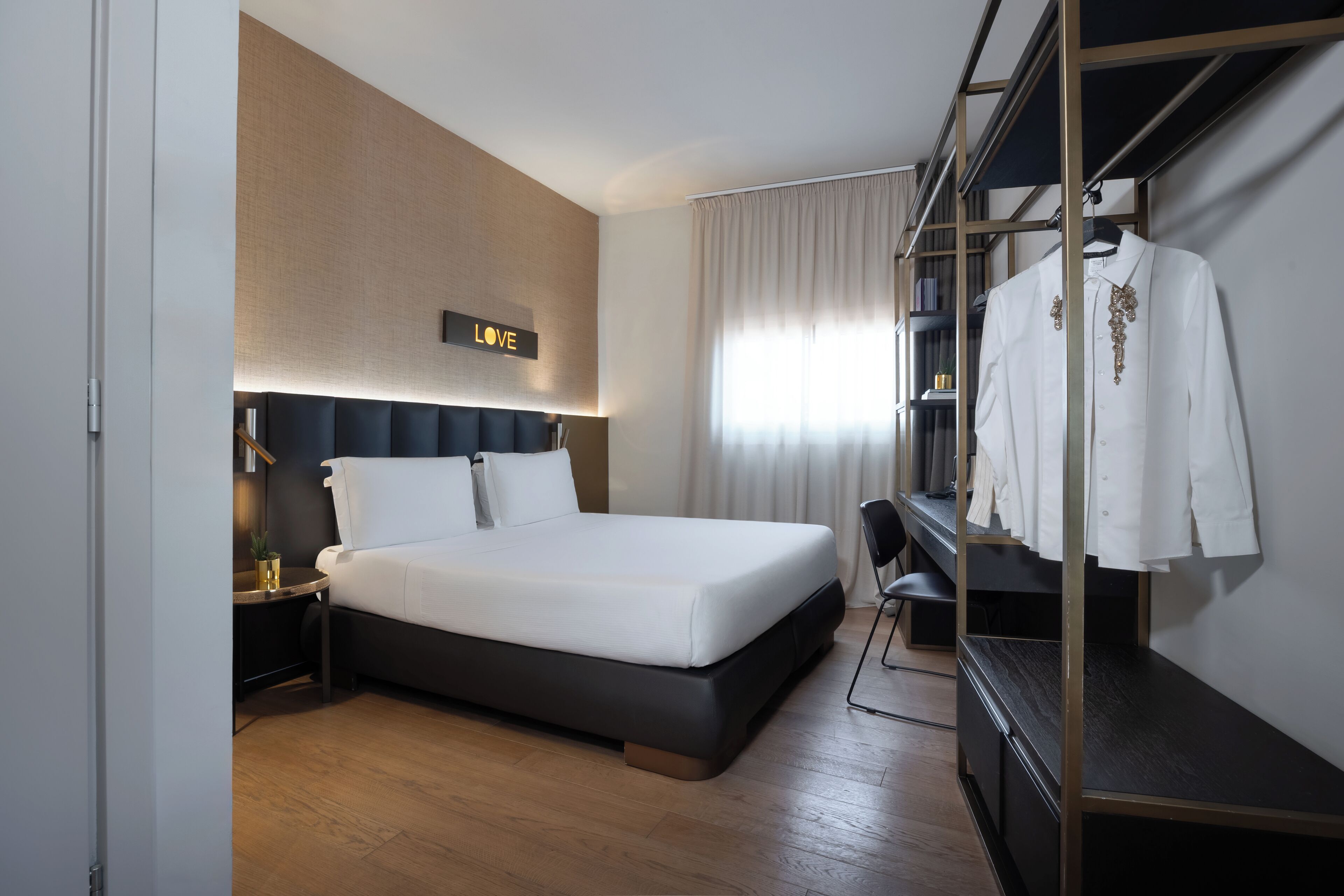 A modern hotel room with a large bed, a wardrobe, a desk, and a wall-mounted sign.