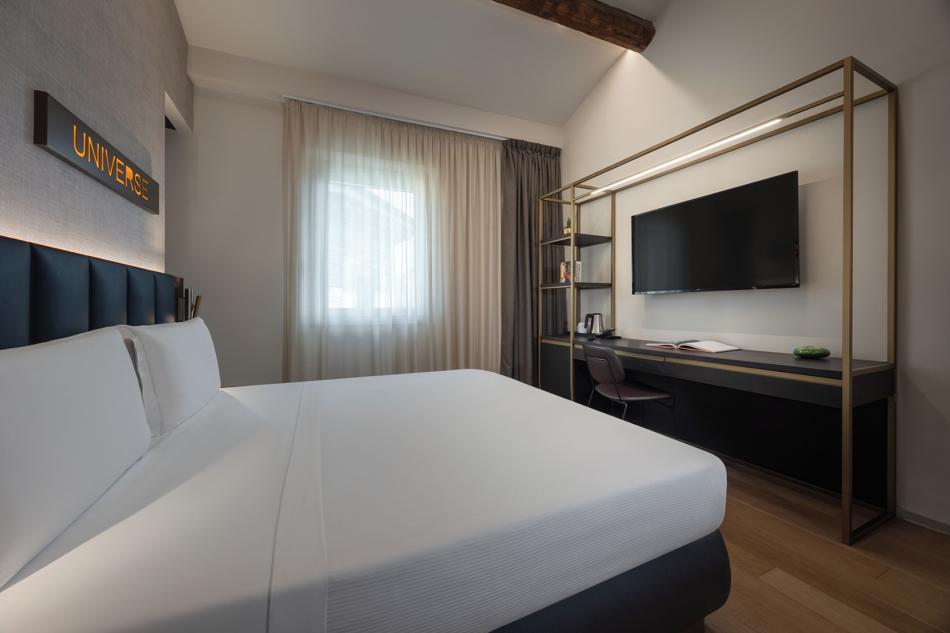 A modern hotel room with a large bed, a desk, a flat-screen TV, and a sign that reads "UNIVERSE".