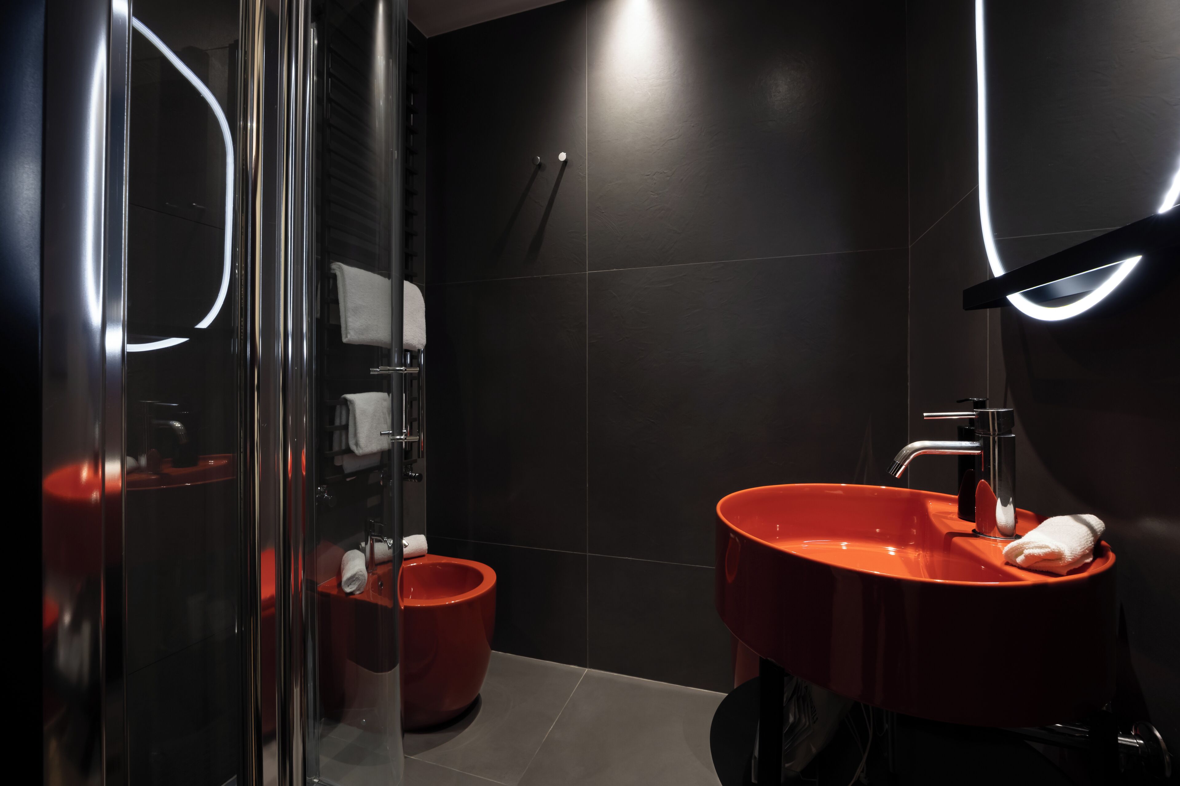Een moderne badkamer met een douchecabine van glas, een rode wastafel en donker betegelde muren.