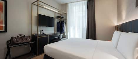 Ein Hotelzimmer mit einem großen Bett, einem Schreibtisch mit Stuhl, einem Kleiderschrank, einem Fernseher und einem Fenster mit Vorhängen.