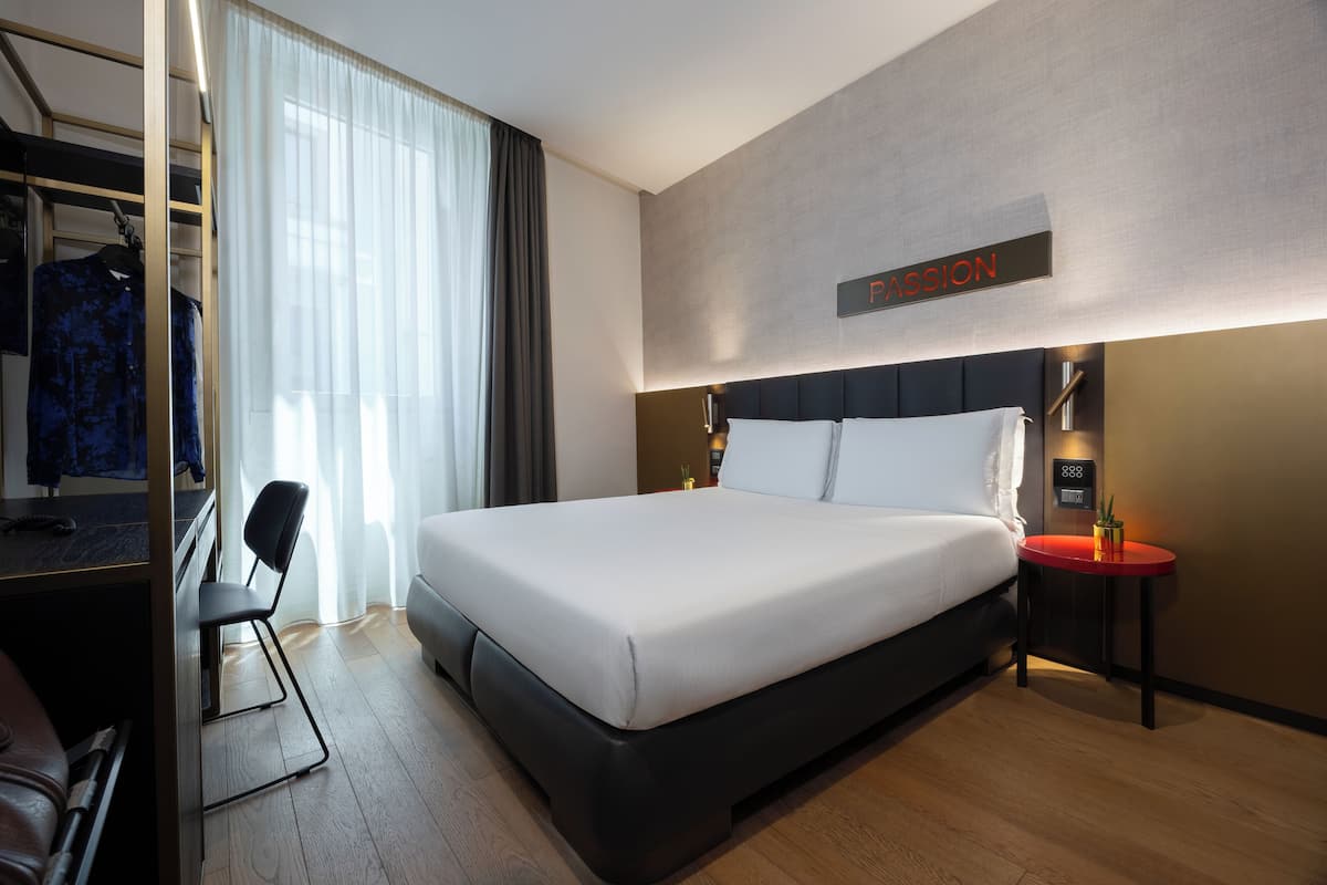 A modern hotel room with a large bed, a desk with a chair, a small table with a plant, and a sign on the wall.
