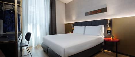 Ein modernes Hotelzimmer mit einem großen Bett, einem Schreibtisch mit Stuhl, einem kleinen Tisch mit Pflanze und einem Schild an der Wand.