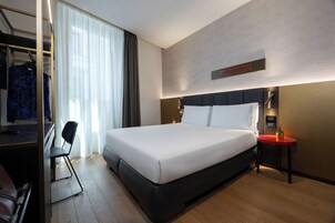 A modern hotel room with a large bed, a desk with a chair, a small table with a plant, and a sign on the wall.