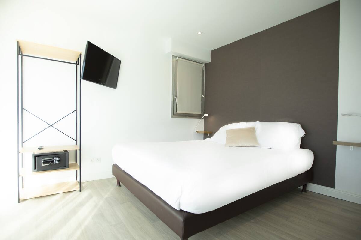 A modern bedroom with a large bed, a flat-screen TV mounted on the wall, a bedside table with a lamp, and a safe.
