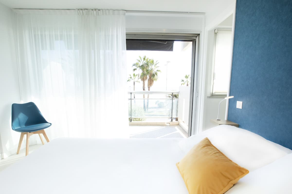 A hotel room with a bed, a blue chair, a balcony with a view of palm trees, and a blue accent wall.