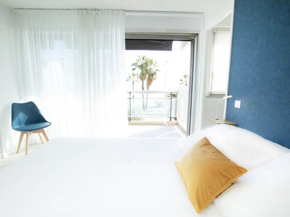 Habitación de hotel con cama, una silla azul, un balcón con vista a palmeras y una pared de acento azul.