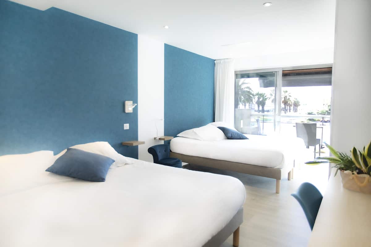 A modern hotel room with two beds, a desk, and a view of the outdoor area.