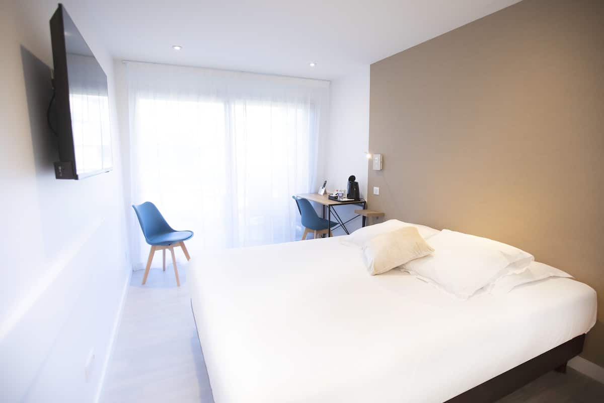 A hotel room with a large bed, a desk with a chair, a television mounted on the wall, and a window with sheer curtains.