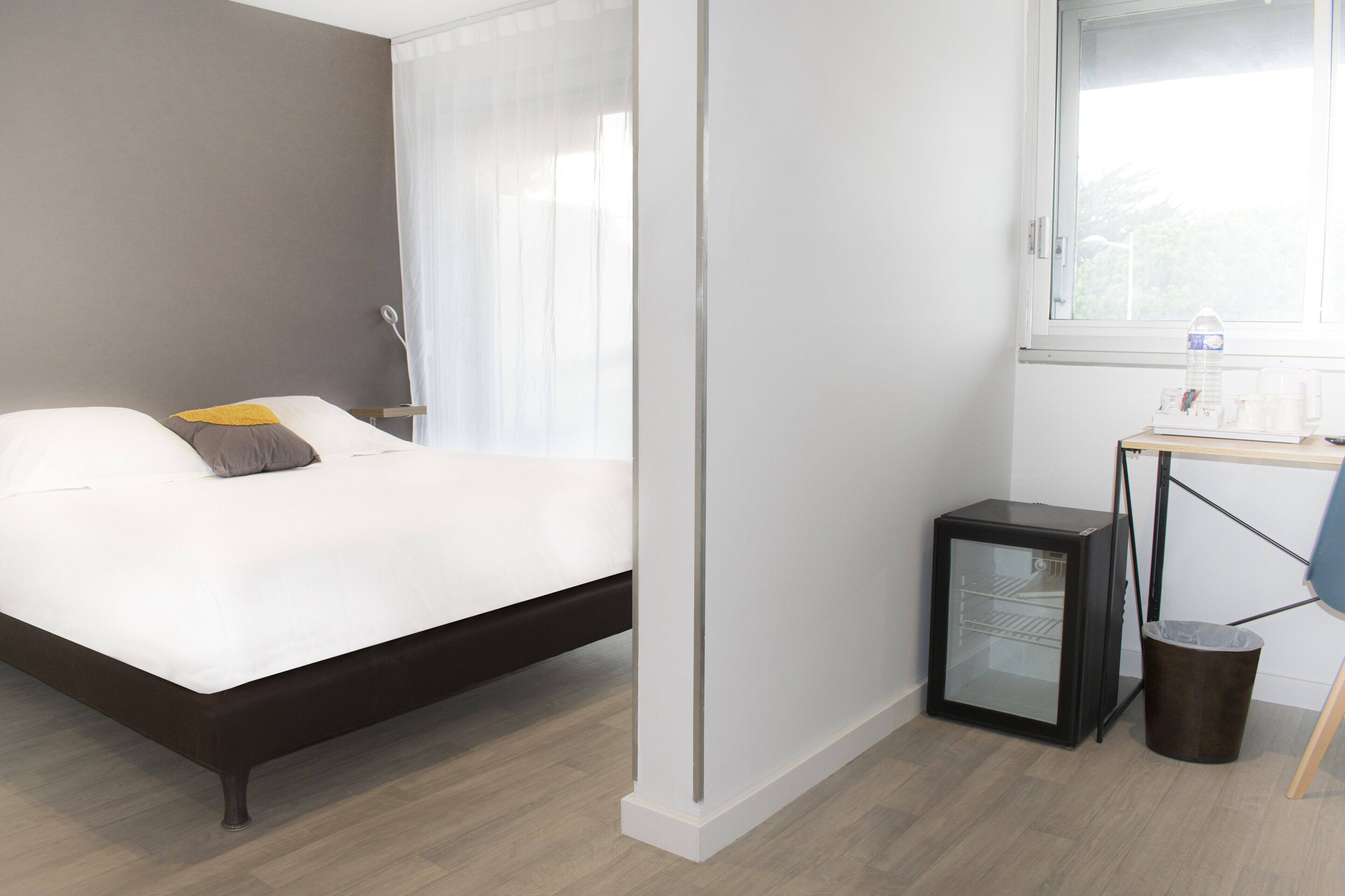 A hotel room with a bed, a desk with a computer, a trash bin, and a window.
