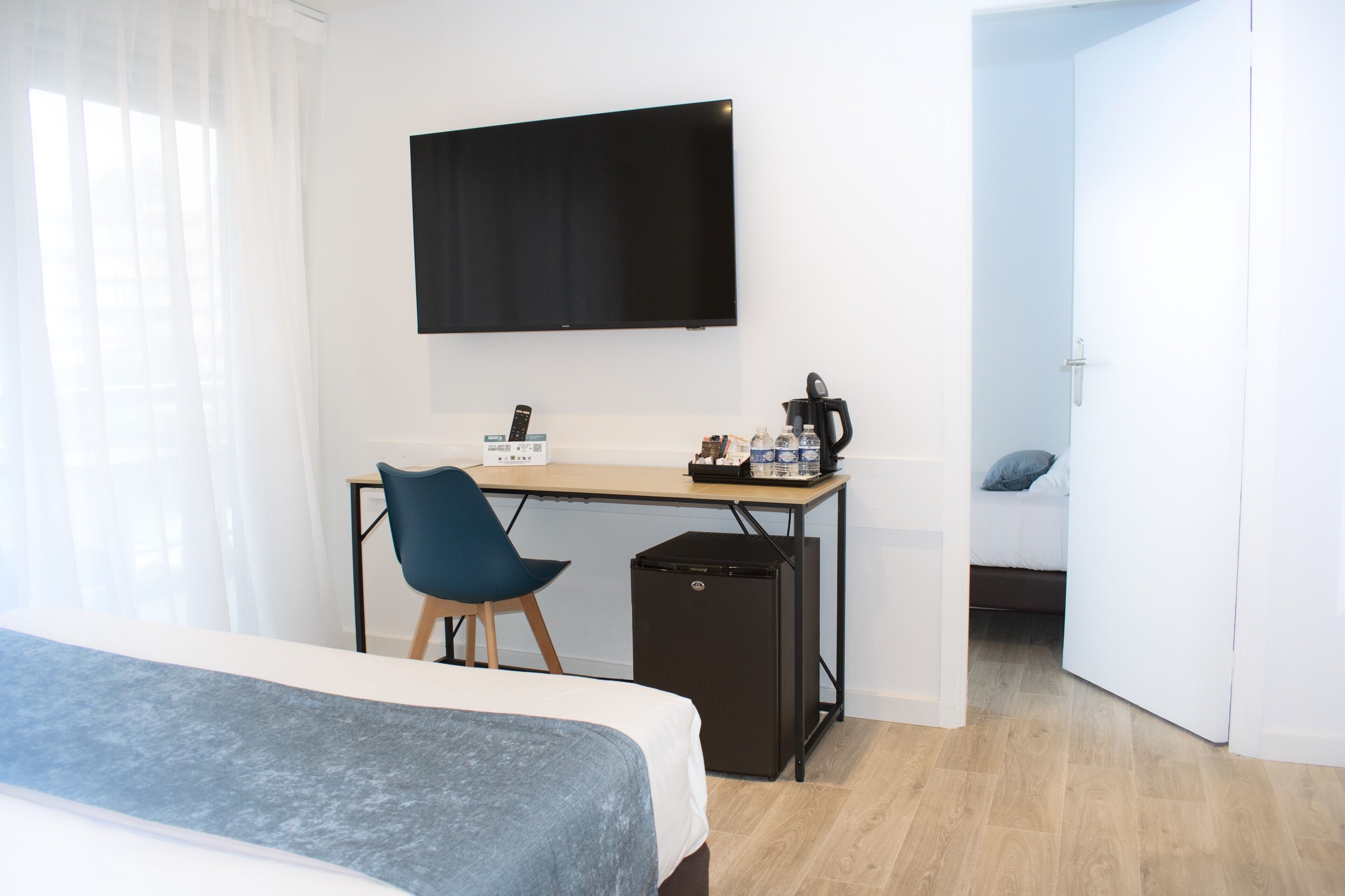 A bedroom with a bed, a desk with a computer, a chair, a TV mounted on the wall, and a door leading to another room.