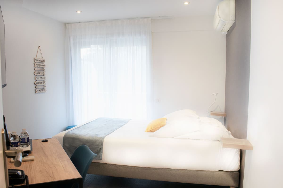 A bedroom with a bed, a desk, a chair, a bedside table, and a window with sheer curtains.