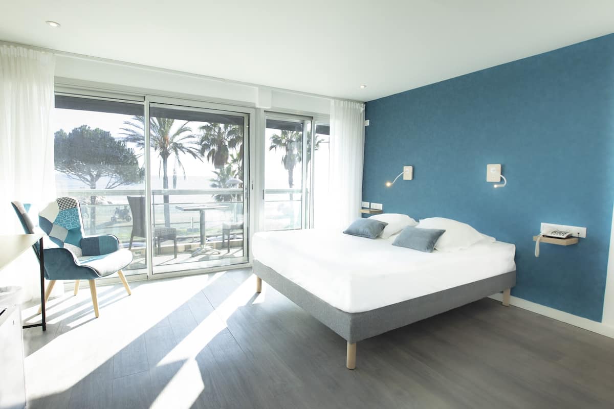 A modern hotel room with a large bed, a balcony with a view of palm trees, and a chair with a view.