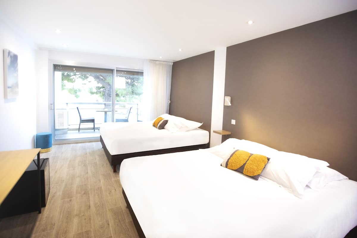 A hotel room with two beds, a wooden floor, a balcony with a table and chairs, and a large window.