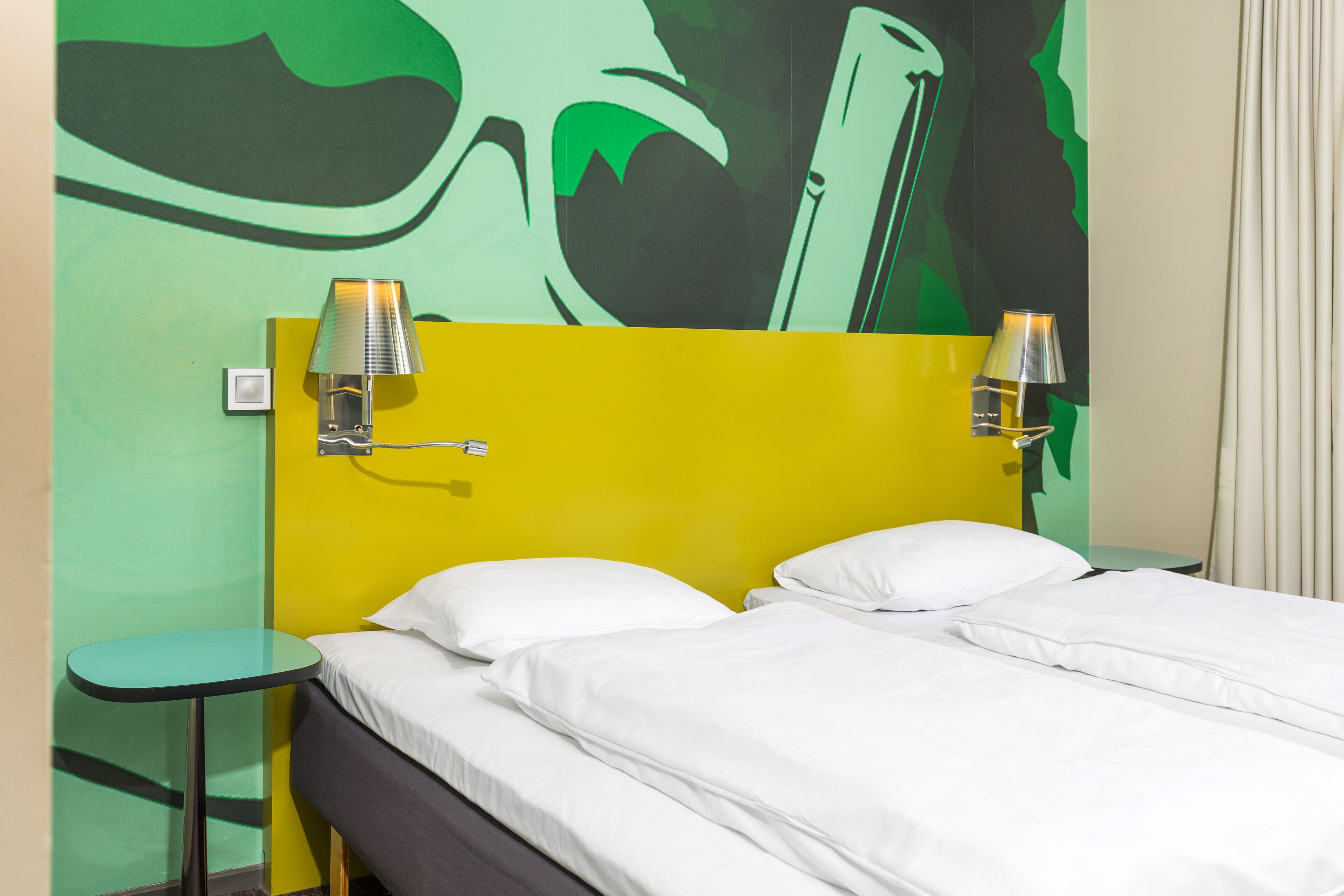 A hotel room with a yellow headboard, two white pillows, a small round table, and two wall-mounted lamps.