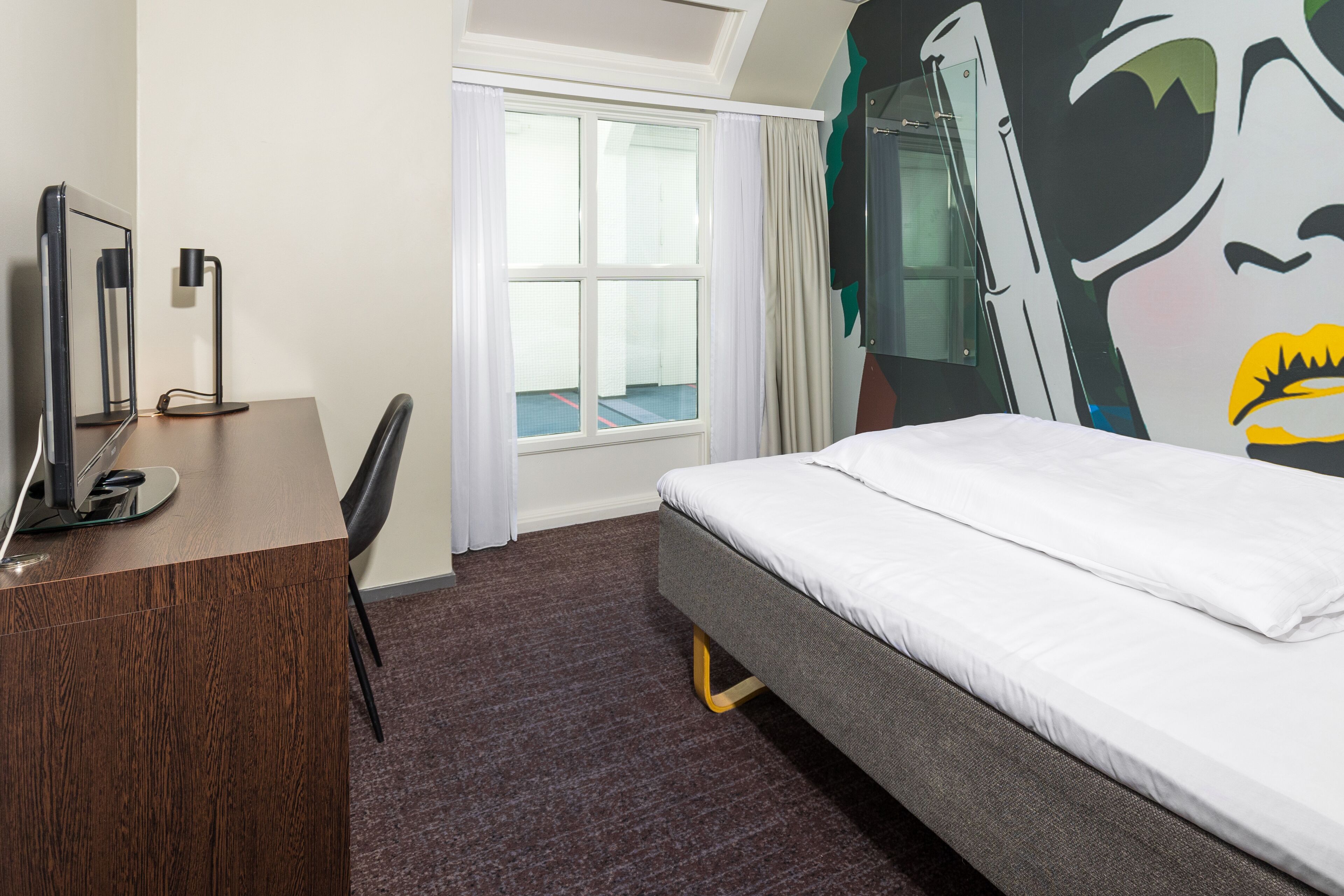 A hotel room with a bed, a desk with a computer, a chair, a window with curtains, and a wall mural.