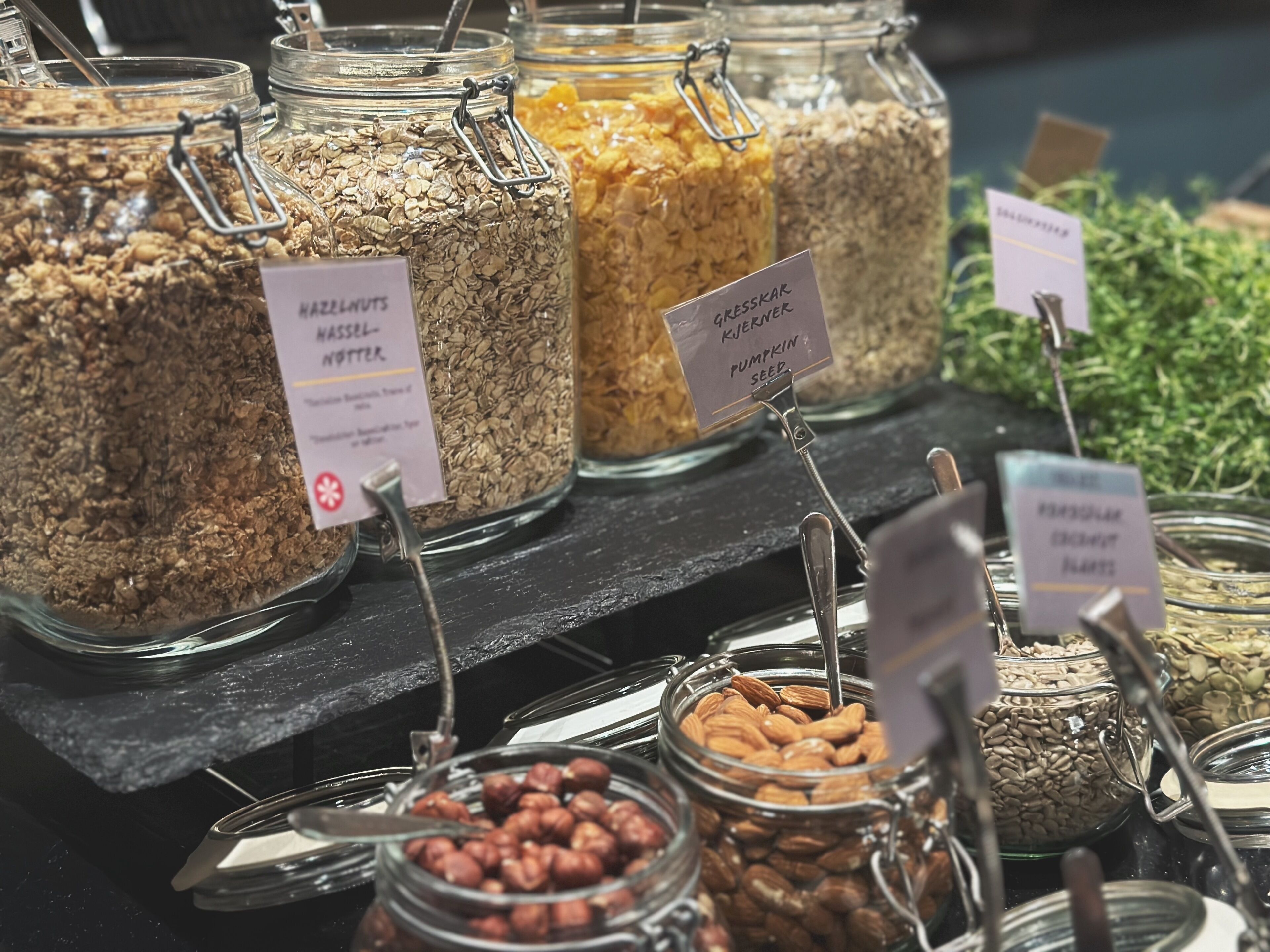 A variety of food items in glass jars with labels, including hazelnuts, oats, and pumpkin seeds.