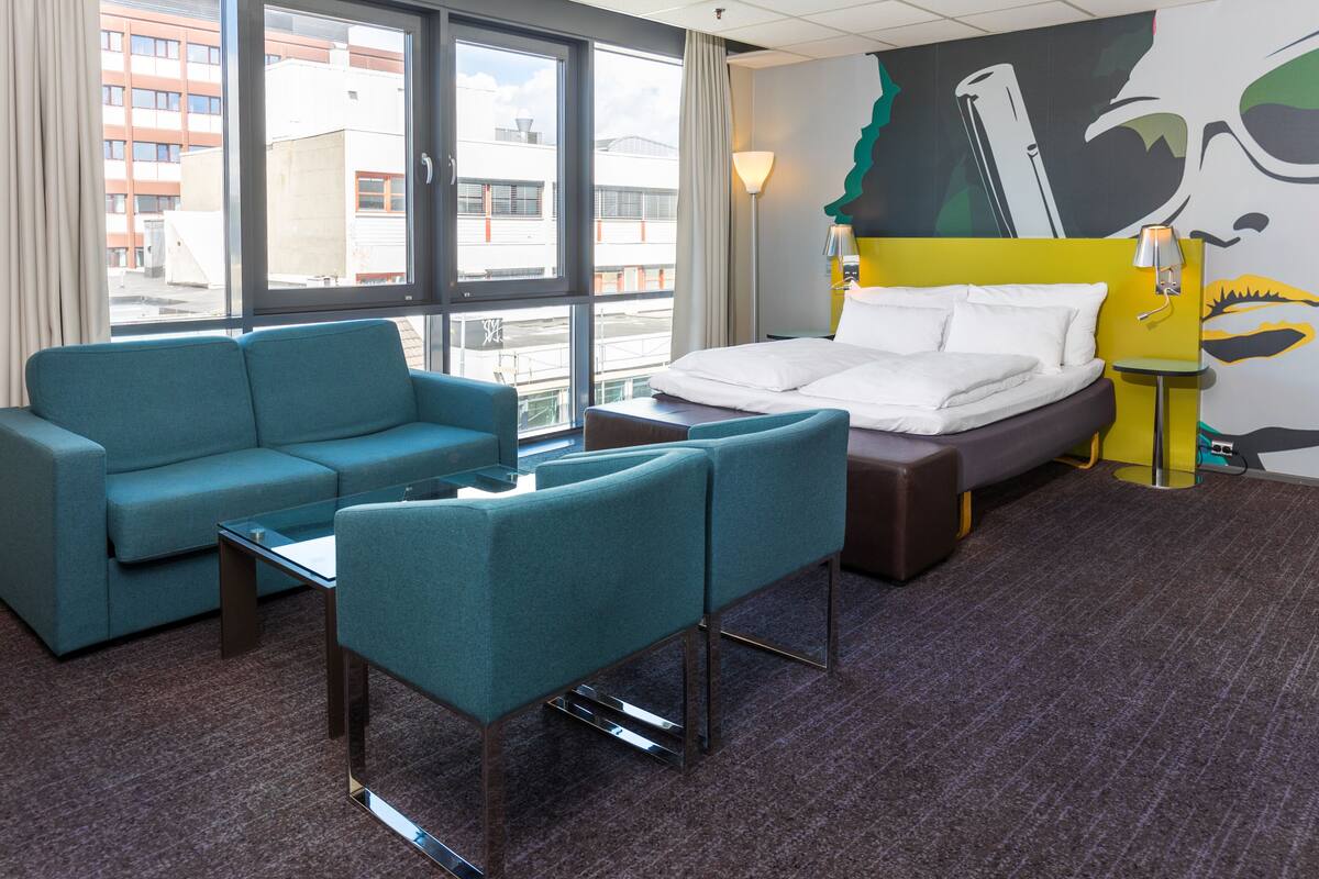 Habitación de hotel con cama, sofá, una mesita y vistas de la ciudad a través de amplias ventanas.