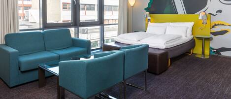 A hotel room with a bed, a sofa, a small table, and a view of the city through large windows.