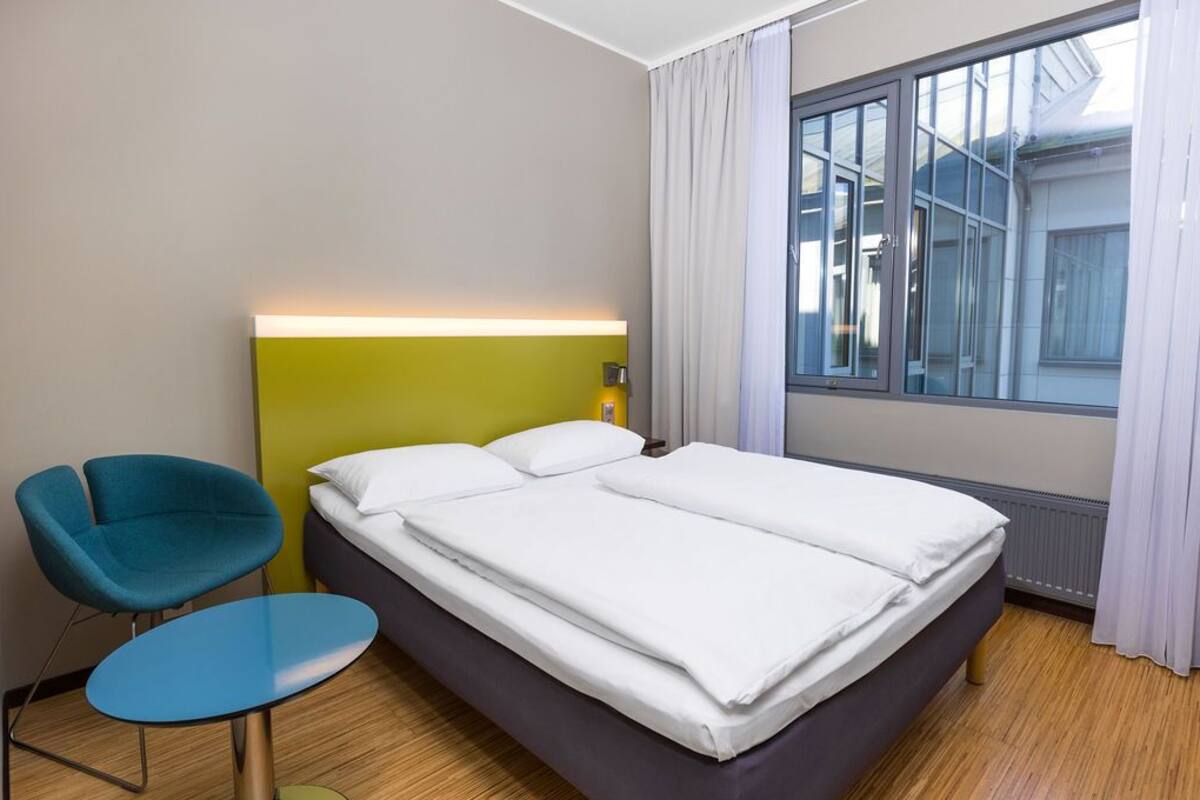Habitación de hotel con cama, una silla, una mesita, ventana con cortinas y una lámpara fijada a la pared.