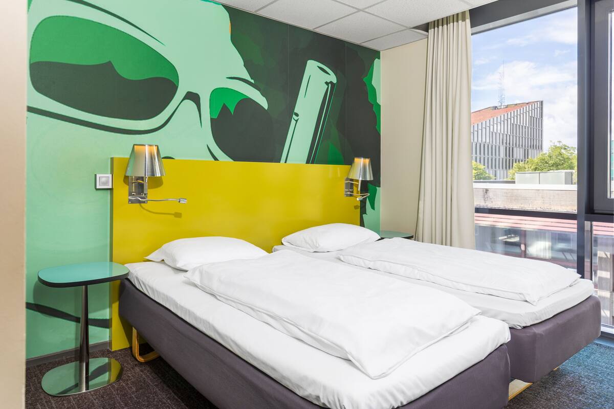 Habitación de hotel con dos camas, un mural en la pared con tonos verde y amarillo, una mesita y una ventana con vistas a edificios.