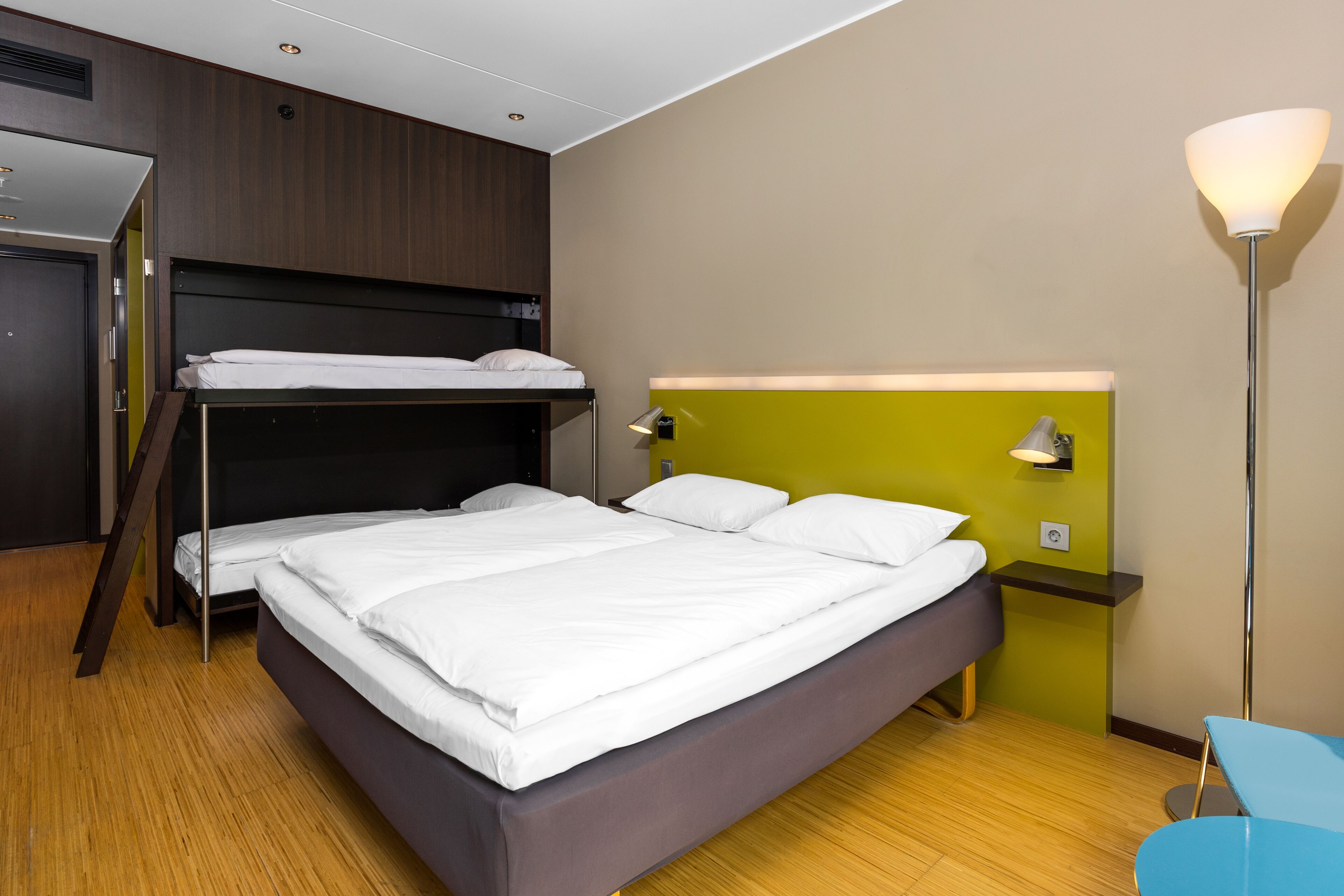 Habitación de hotel con una cama grande, una cama litera más pequeña, un escritorio, una silla y una lámpara de pie.
