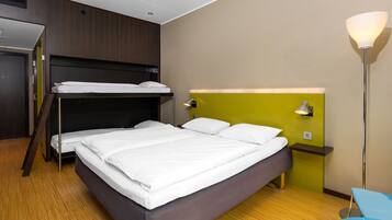 Quarto de hotel com uma cama grande, uma cama beliche menor, uma escrivaninha, uma cadeira e um abajur.