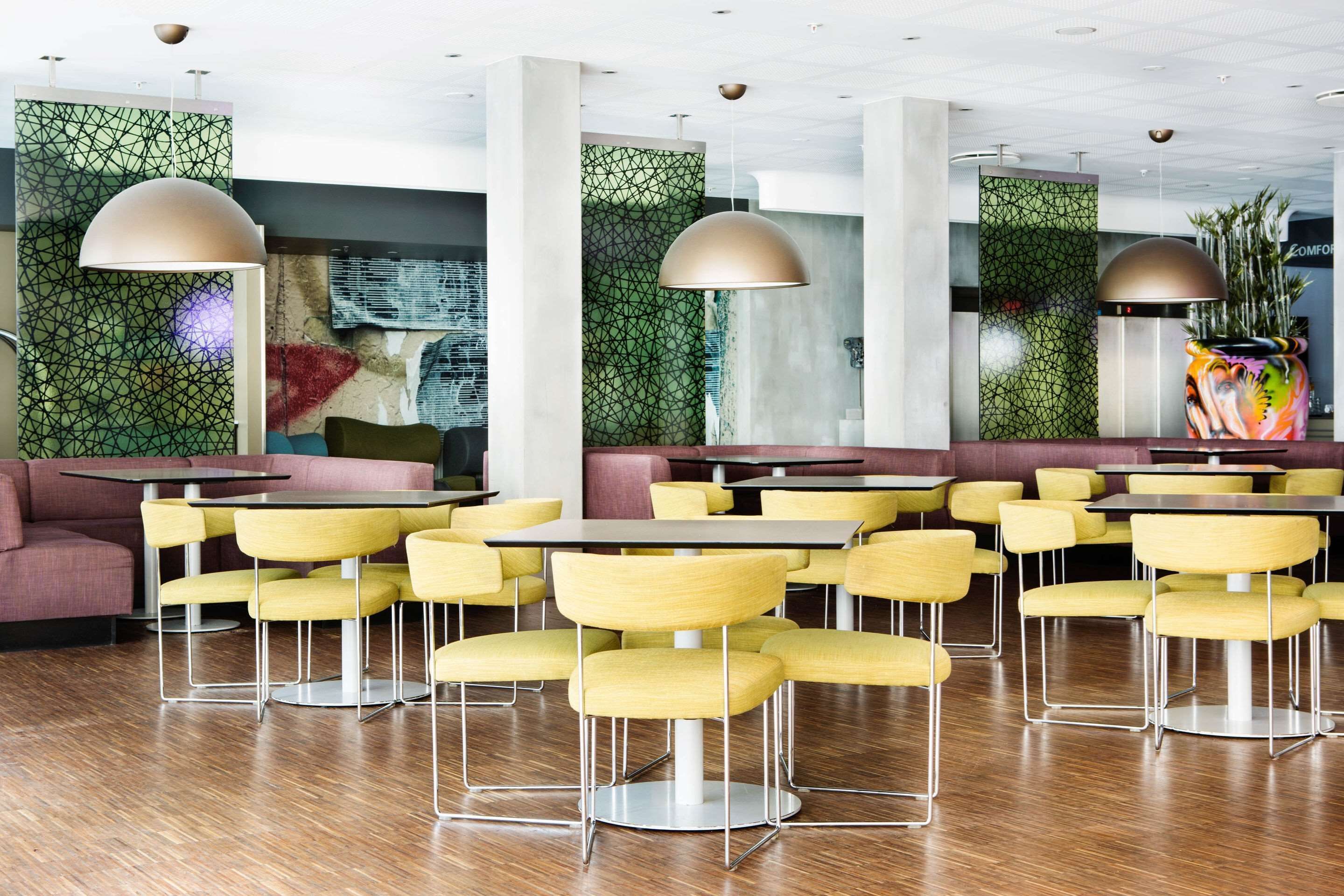A modern restaurant with yellow chairs and tables, green patterned walls, and large pendant lights.