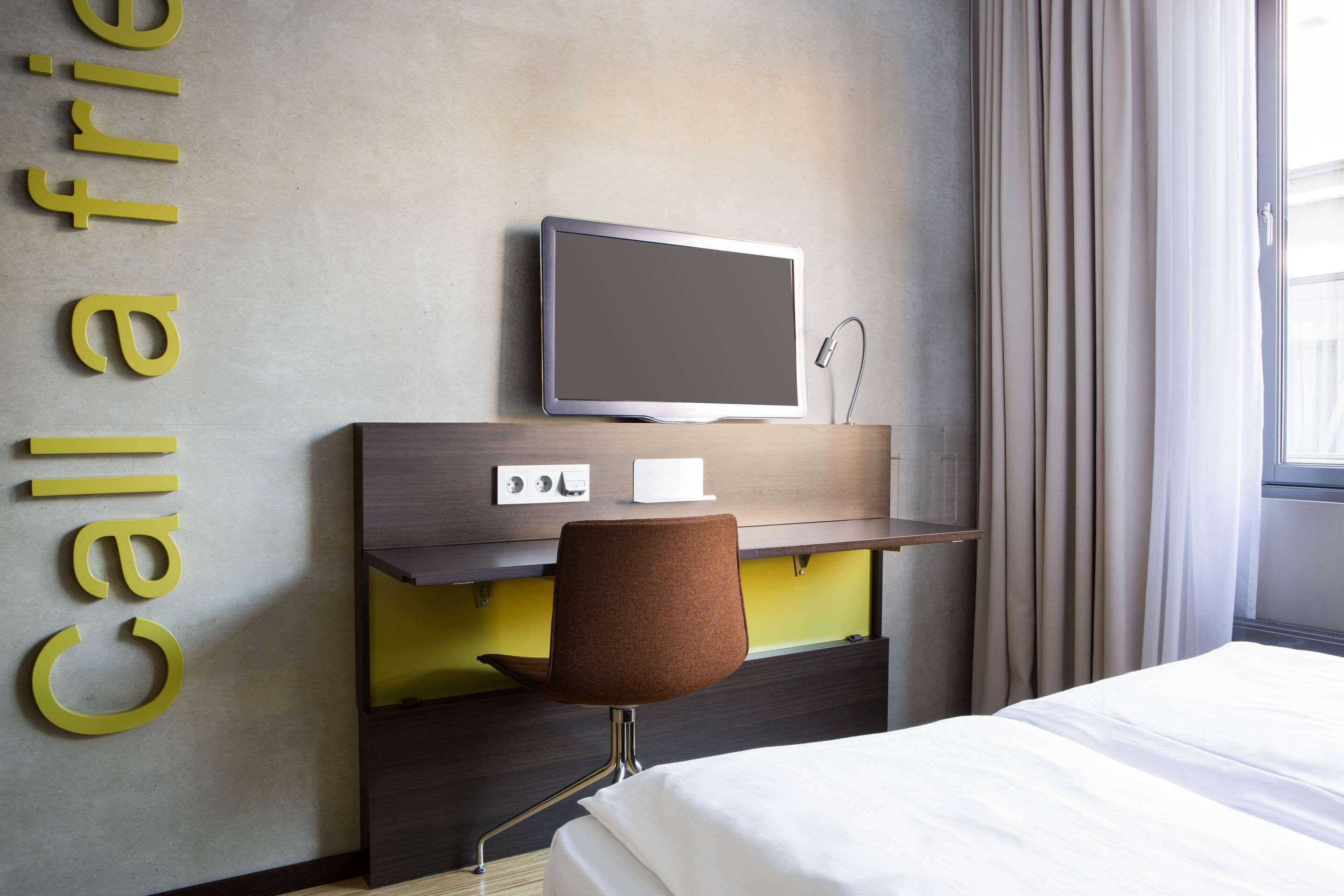 A hotel room with a bed, a desk with a chair, a television, and a wall-mounted control panel.