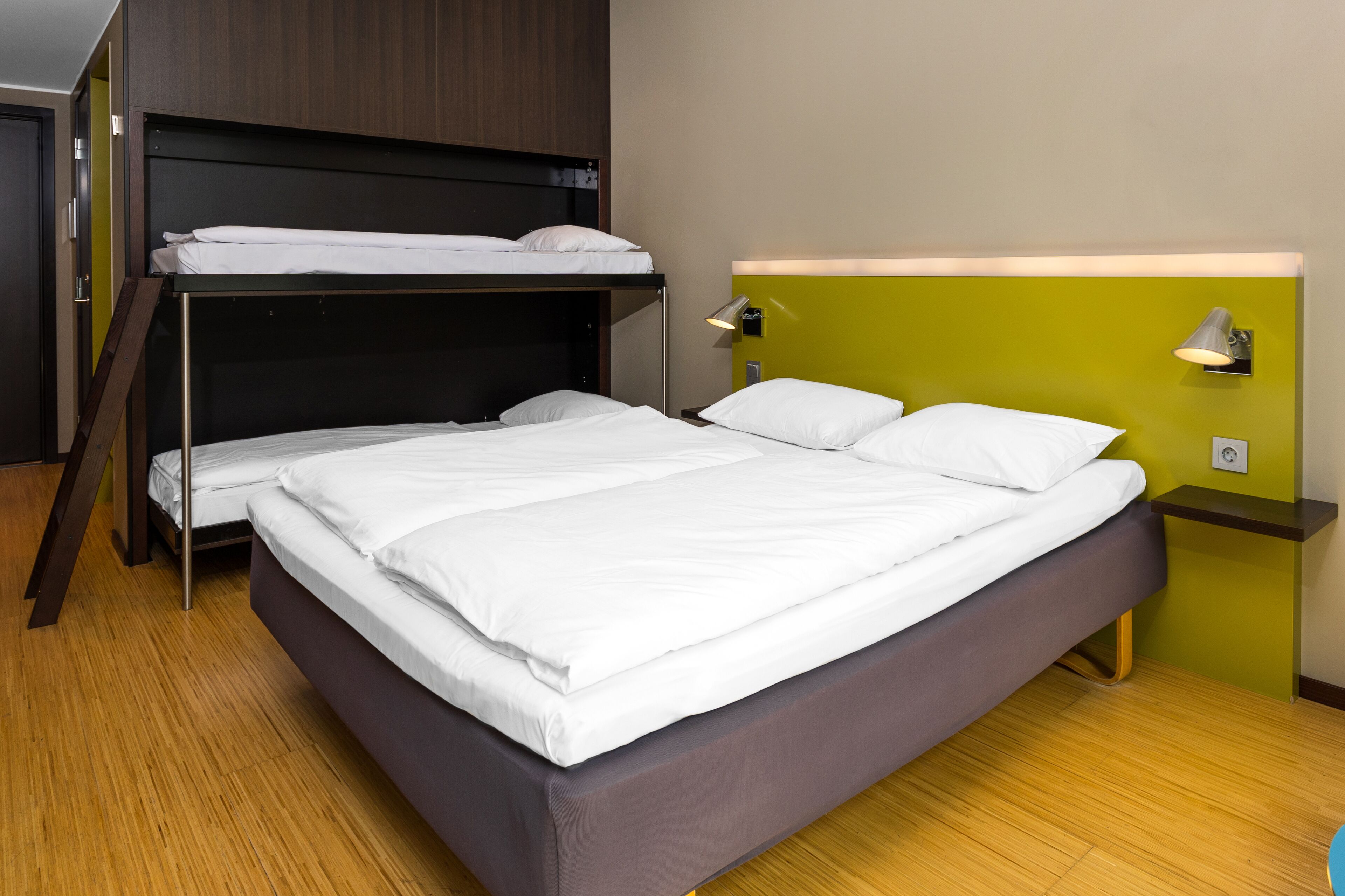 A room with a bunk bed, a single bed, a bedside table with a lamp, and a wall socket.