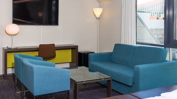 Une chambre d’hôtel moderne avec un canapé bleu, une table basse en verre, un fauteuil marron et un téléviseur à écran plat fixé au mur.