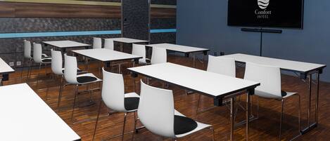 A conference room with white tables and chairs, a wooden floor, and a TV screen displaying the Comfort Hotel logo.