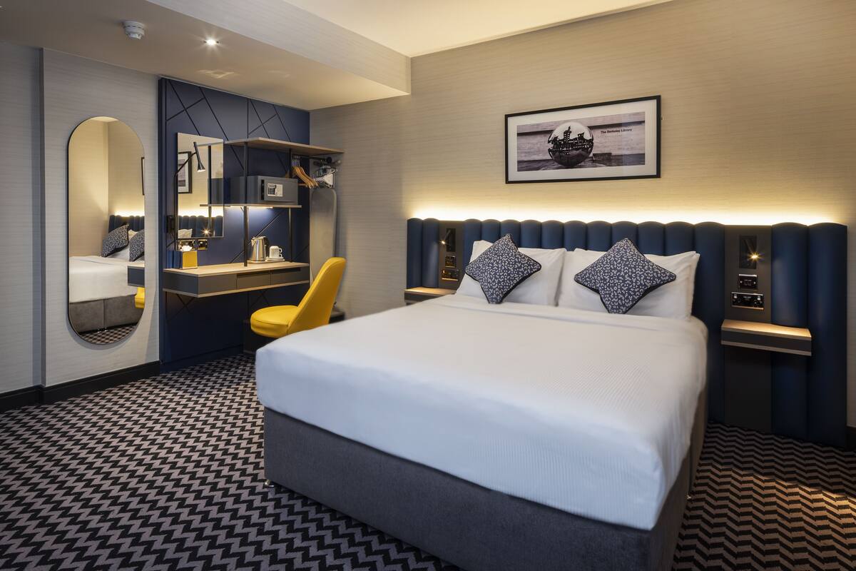 A modern hotel room with a large bed, a desk with a chair, a mirror, and a framed picture on the wall.