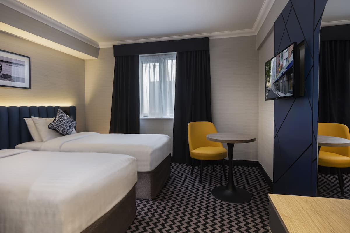 A hotel room with two beds, a small table, two chairs, a TV, and a window with curtains.
