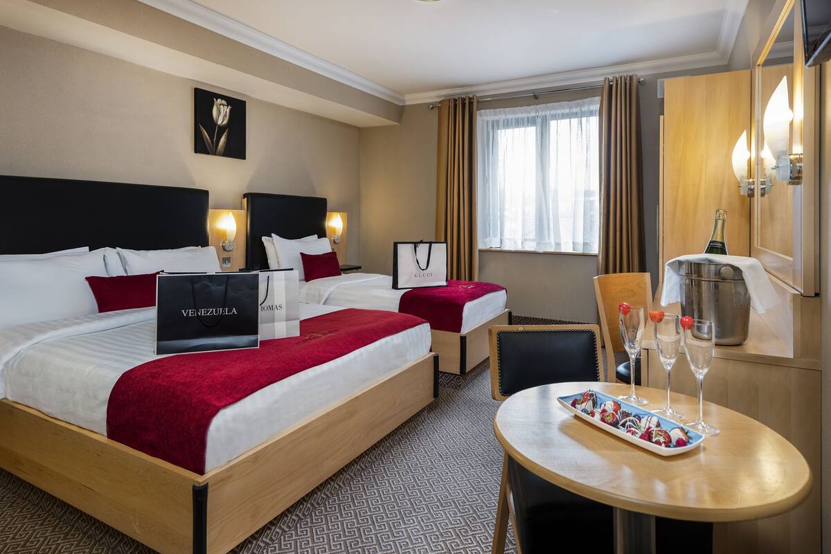 A hotel room with two beds, a dining table with a fruit platter, a minibar, and a bottle of champagne.