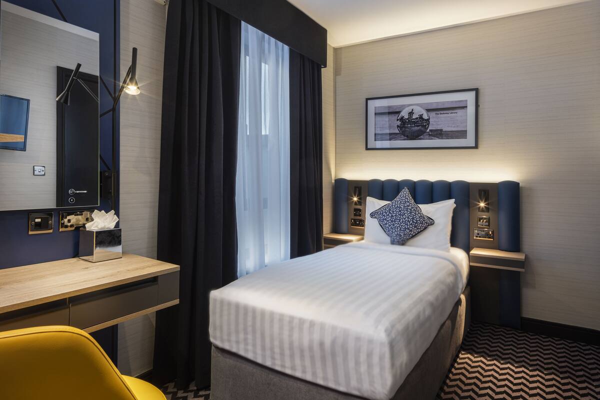 A hotel room with a bed, a desk, a chair, a nightstand, and a wall-mounted lamp.